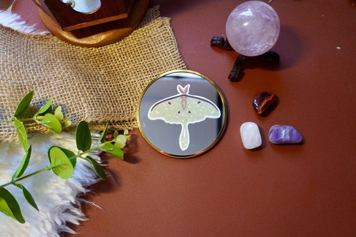 The image shows a sticker of a luna moth on a circle mirror. The moth is light green with intricate white star, moon, and dot designs on its wings, along with dark purple accents and a brown body. The sticker is die-cut to follow the shape of the moth, giving it a distinct and whimsical appearance. 