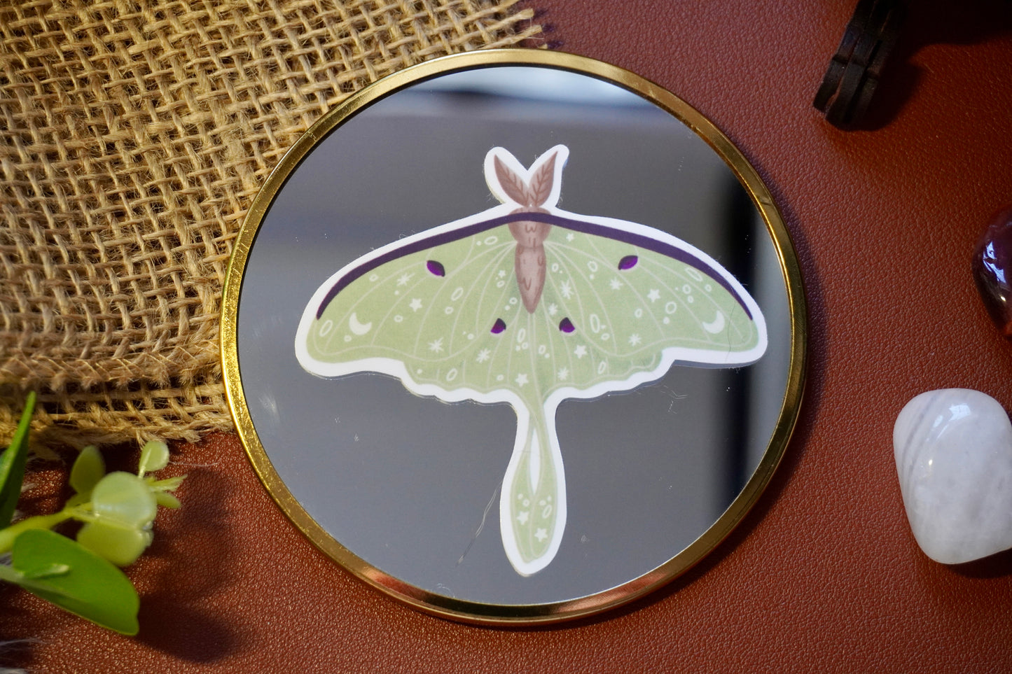 The image shows a sticker of a luna moth on a circle mirror. The moth is light green with intricate white star, moon, and dot designs on its wings, along with dark purple accents and a brown body. The sticker is die-cut to follow the shape of the moth, giving it a distinct and whimsical appearance. 