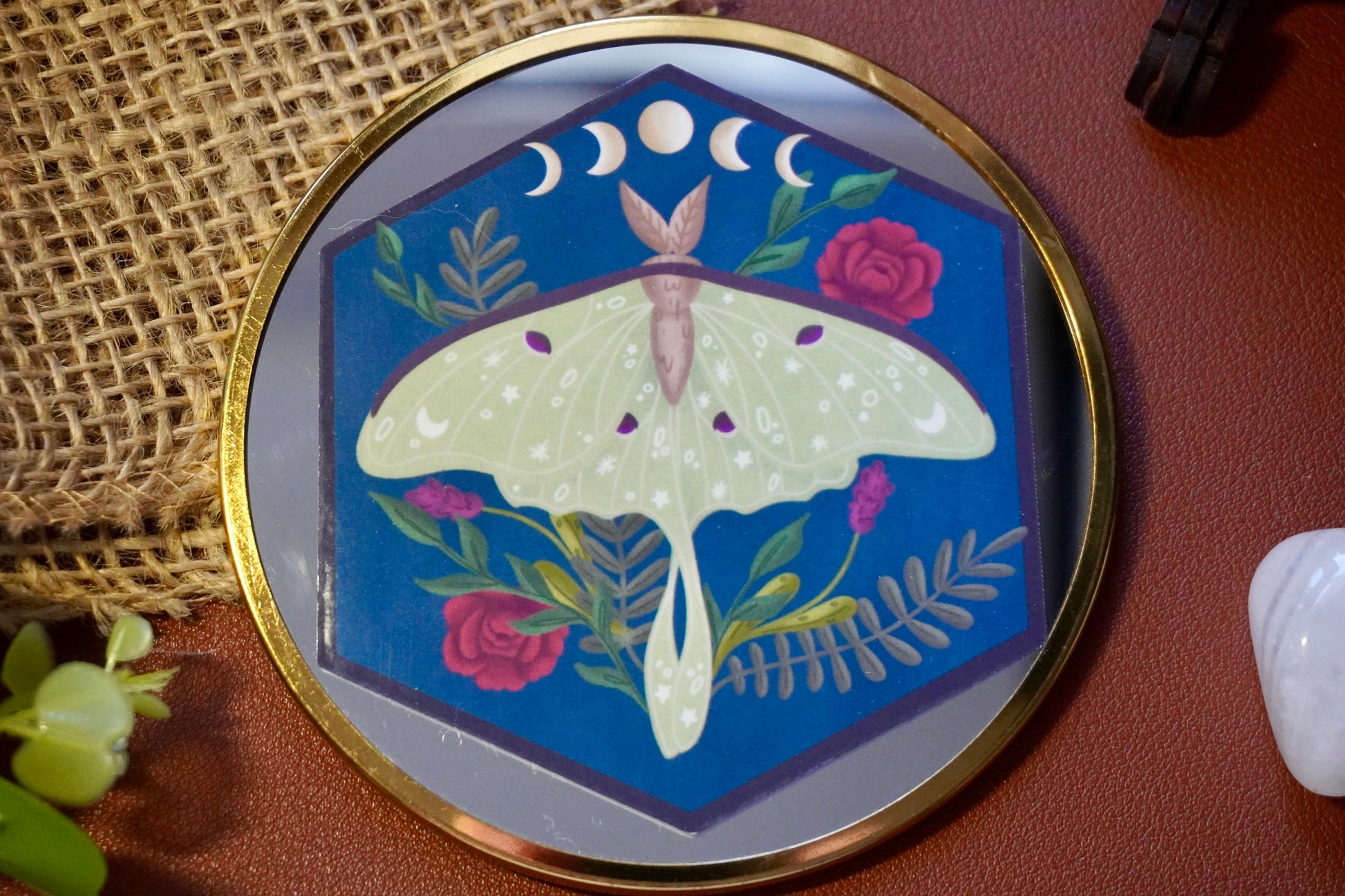 The image shows a hexagon-shaped sticker. The design features a green luna moth with decorative star and dot patterns on its wings, surrounded by various plants including red roses and ferns. Above the moth, a lunar phase motif is displayed, showing the progression of the moon from waxing crescent to full and waning crescent. The background is a deep blue, enhancing the contrast with the soft green moth and vibrant plant colors. The overall style is whimsical and nature-inspired.