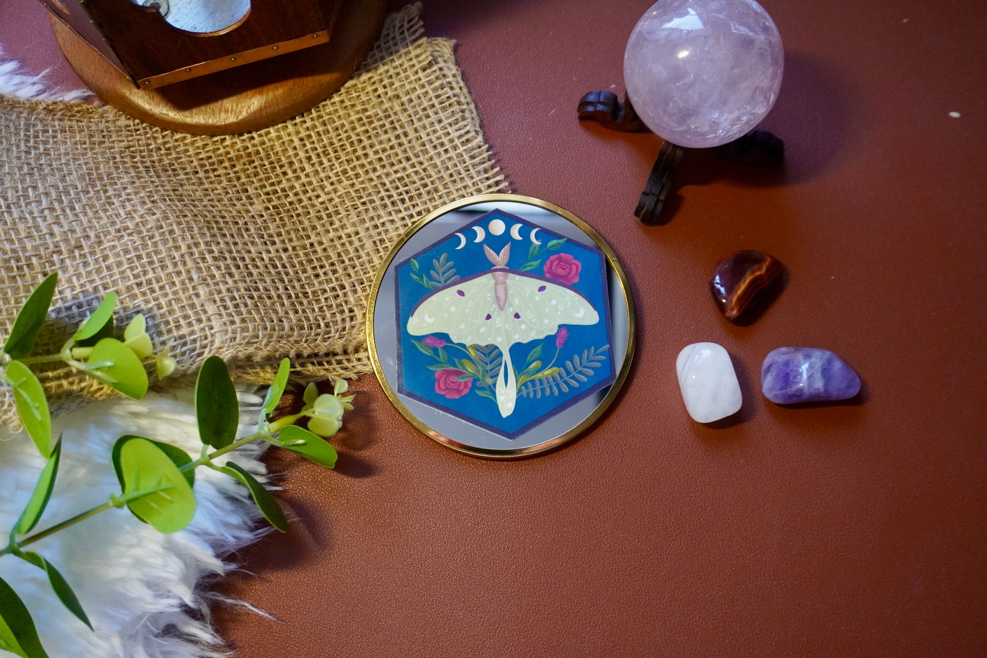 The image shows a hexagon-shaped sticker. The design features a green luna moth with decorative star and dot patterns on its wings, surrounded by various plants including red roses and ferns. Above the moth, a lunar phase motif is displayed, showing the progression of the moon from waxing crescent to full and waning crescent. The background is a deep blue, enhancing the contrast with the soft green moth and vibrant plant colors. The overall style is whimsical and nature-inspired.