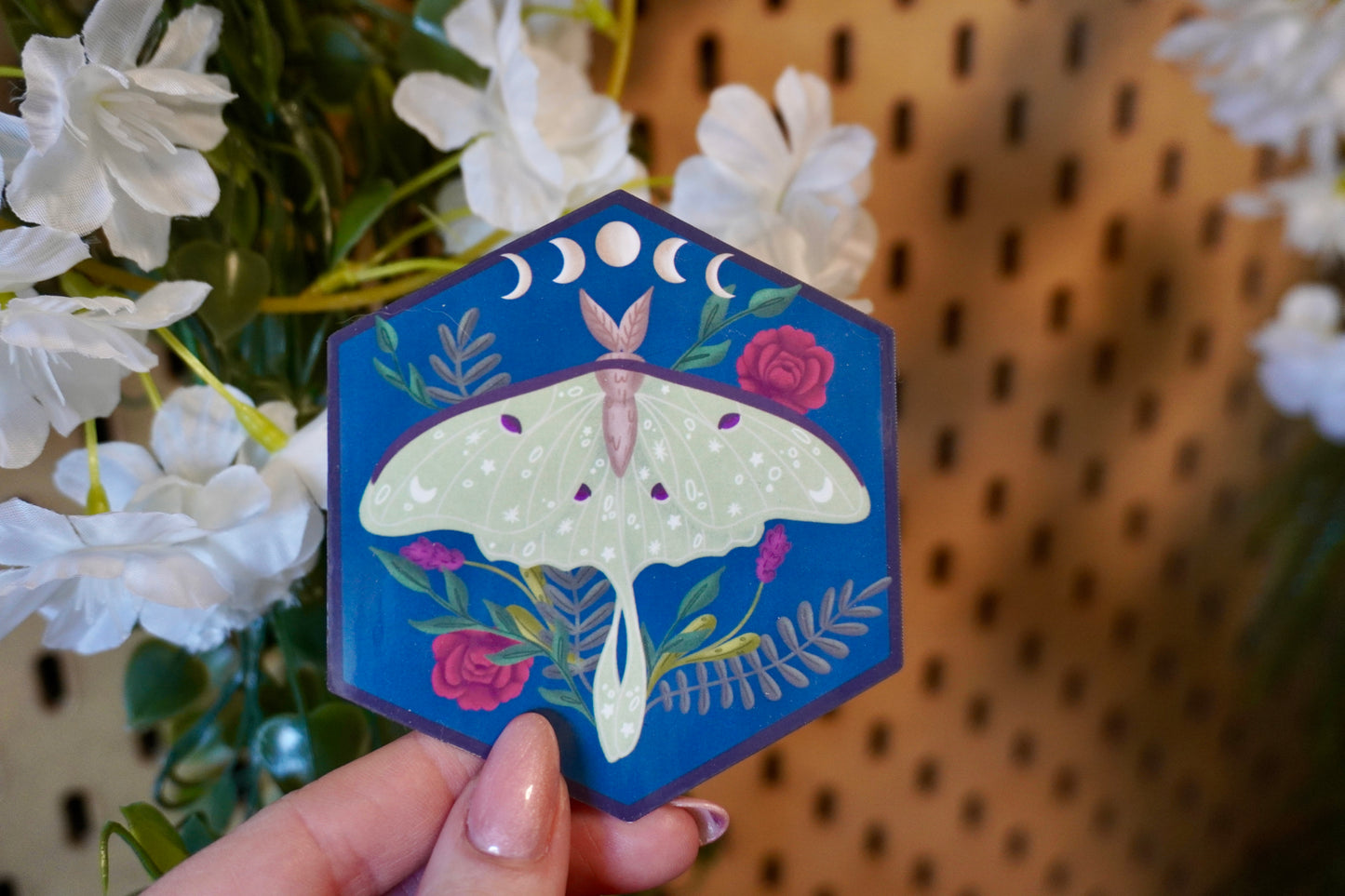 The image shows a hexagon-shaped sticker being held up in front of some artificial white flowers. The design features a green luna moth with decorative star and dot patterns on its wings, surrounded by various plants including red roses and ferns. Above the moth, a lunar phase motif is displayed, showing the progression of the moon from waxing crescent to full and waning crescent. The background is a deep blue, enhancing the contrast with the soft green moth and vibrant plant colors. 