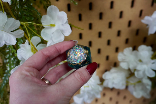Hand holding a large, handcrafted resin D20 die with a shimmering iridescent core and gold accents, set against a backdrop of white artificial flowers and a pegboard wall.